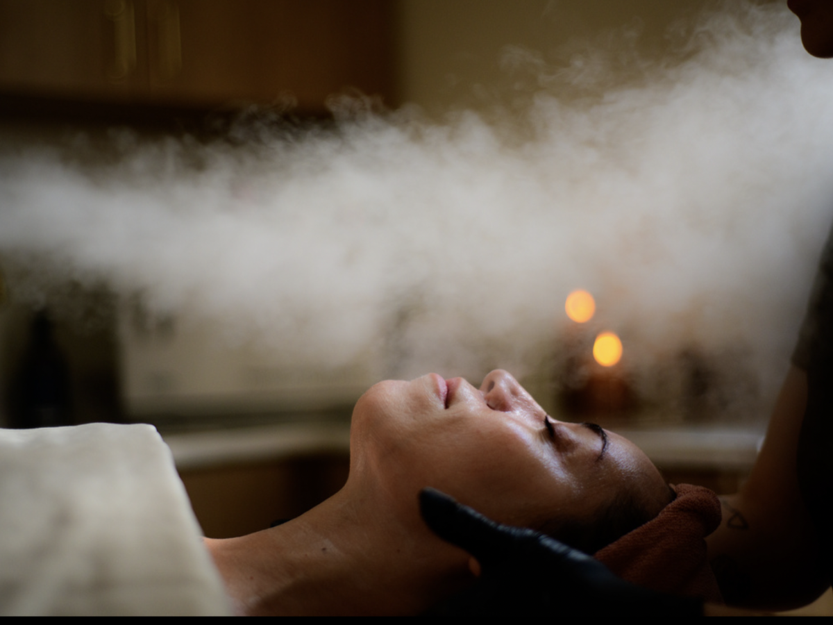 A person is lying down with steam directed at their face in a spa setting. Candles are blurred in the background, creating a calming ambiance.