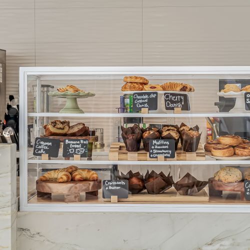A glass display case with assorted pastries and cakes, labeled with small chalk signs; a coffee machine is visible to the left.