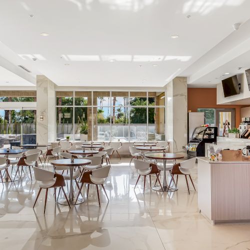 A bright, modern cafe or dining area with white tiled floors, wooden chairs, round tables, large windows, and a cafe counter on the right.