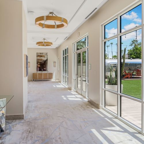 Bright sunlit glass-walled corridor with marble floors, modern lamps, and a view into a green backyard through large windows.