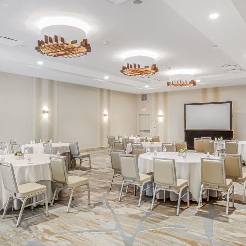 A bright banquet room with round tables, beige chairs, white tablecloths, wall lights, and a large screen at the front, ready for a conference.