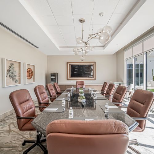 A modern conference room with a long glass table, twelve brown leather chairs, chic chandelier, marble floor, and large windows letting in natural light.