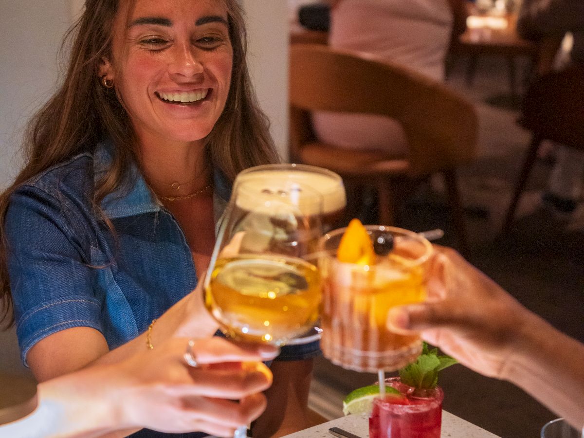 A smiling woman in a denim shirt toasting drinks with friends at a restaurant, colorful cocktails and a lively evening vibe, cheers.