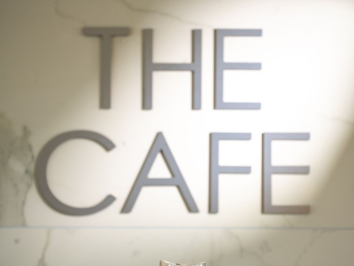 A hand holds a iced coffee in front of a wall with the words &ldquo;THE CAFE&rdquo; lit up, background marble texture, soft spotlight.