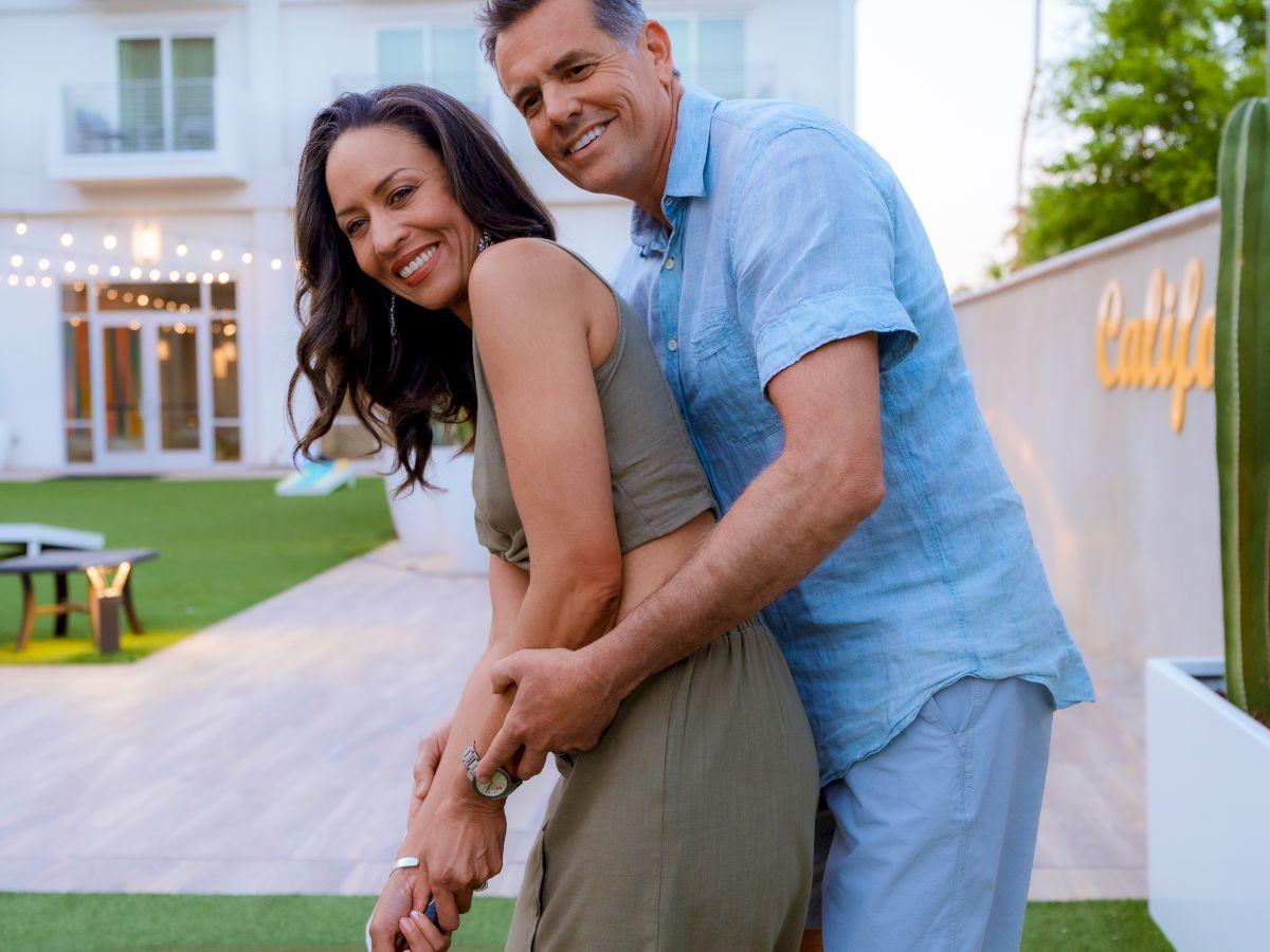 A couple is playing mini-golf together, smiling and embracing, with a building in the background and a green lawn underfoot.