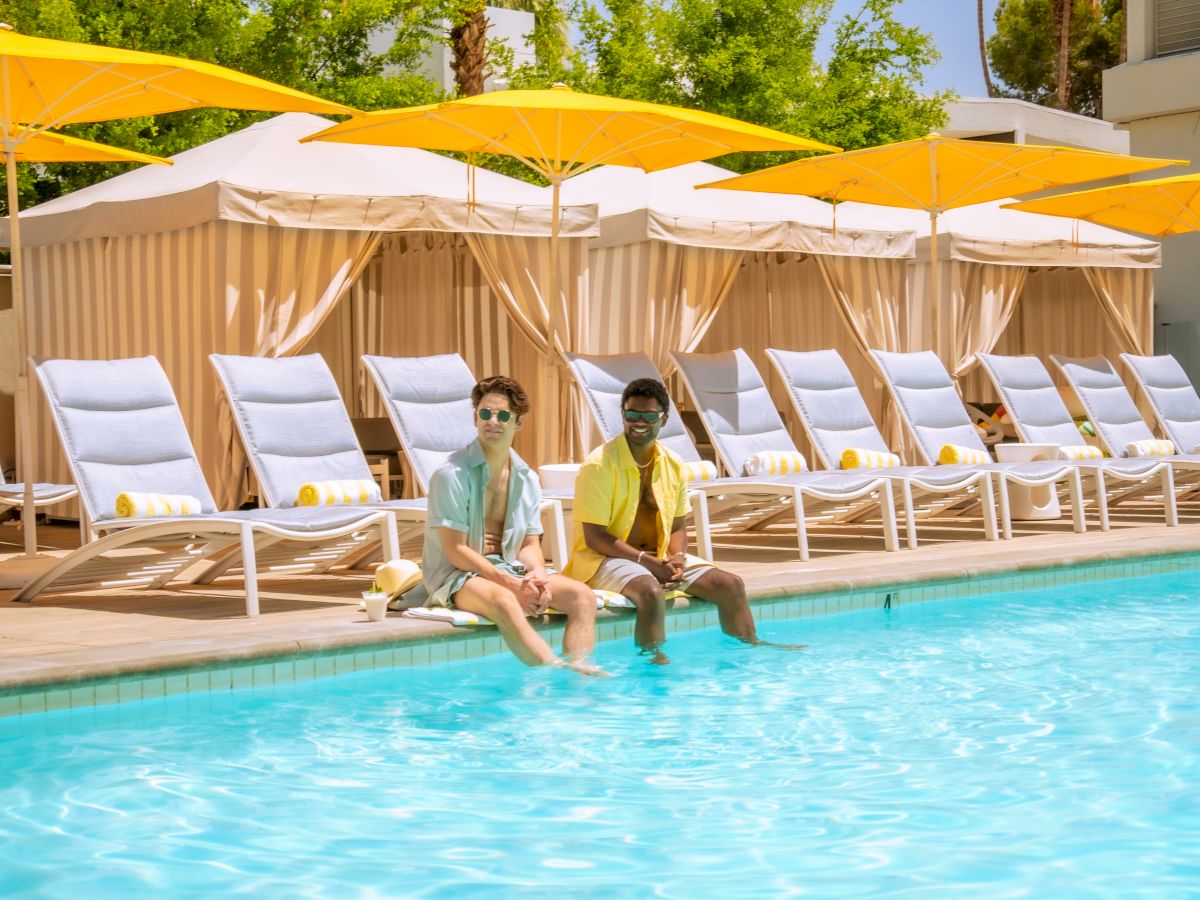 Two people sitting by a pool with lounge chairs and cabanas, surrounded by palm trees, under a clear blue sky.