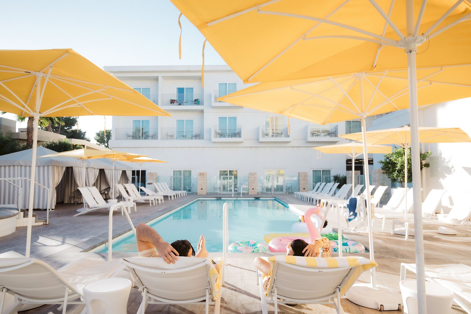 A sunny poolside scene with yellow umbrellas, lounge chairs, and people relaxing near a swimming pool at a hotel or resort.