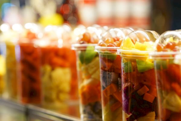 The image shows clear cups filled with assorted fresh fruit pieces, lined up on a counter.