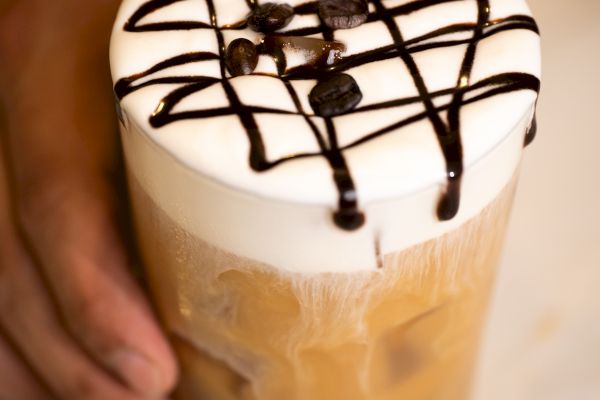 A hand holds an iced coffee topped with foam, drizzled with chocolate syrup in a lattice pattern, and garnished with coffee beans.