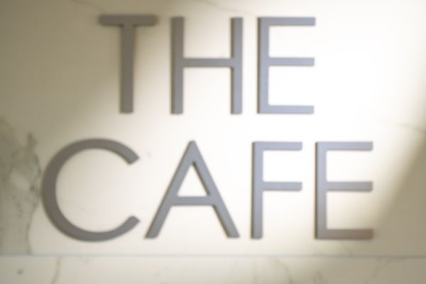 A hand holds an iced coffee with whipped cream in front of a wall with the text "THE CAFE" illuminated by light.