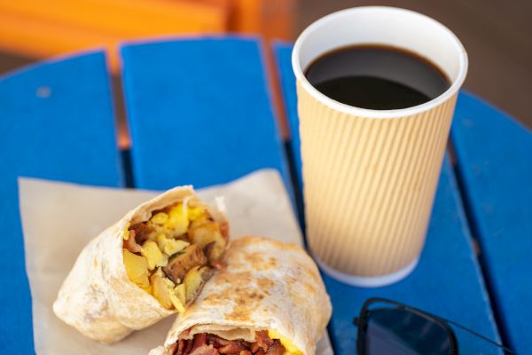 A breakfast burrito cut in half on paper, a cup of black coffee, and sunglasses on a blue table.