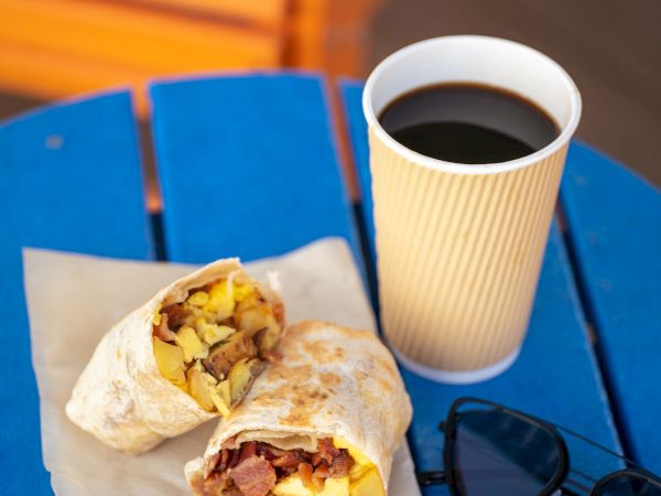 A breakfast burrito cut in half on paper, a cup of black coffee, and sunglasses on a blue table.