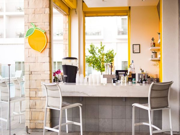 The image shows a bar with two stools, a lemon sign, various drinks, and a bright, inviting atmosphere.