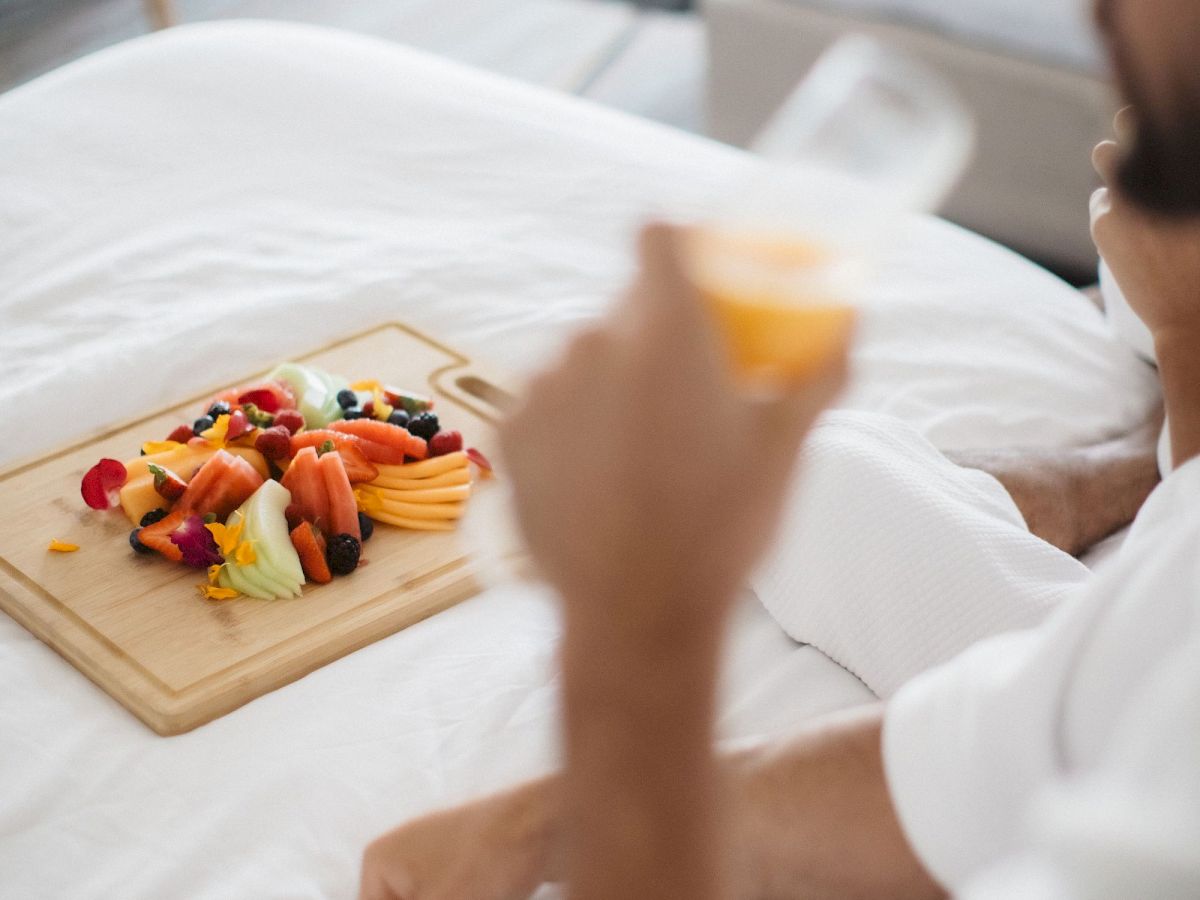 A person is sitting on a bed holding a drink, with a wooden board of assorted fruits nearby.