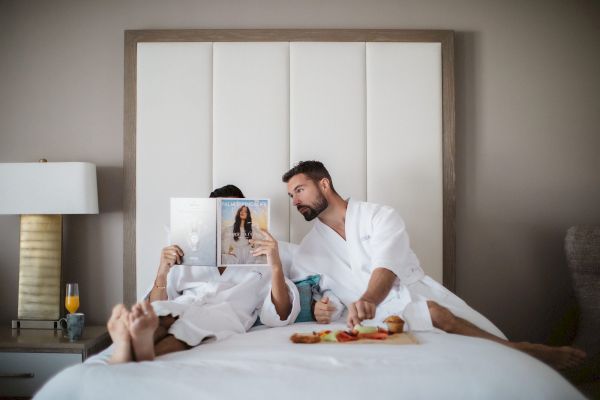 Two people in white robes relax on a bed; one reads a magazine while the other reaches for food on a tray.