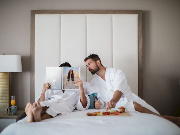 Two people in white robes relax on a bed; one reads a magazine while the other reaches for food on a tray.
