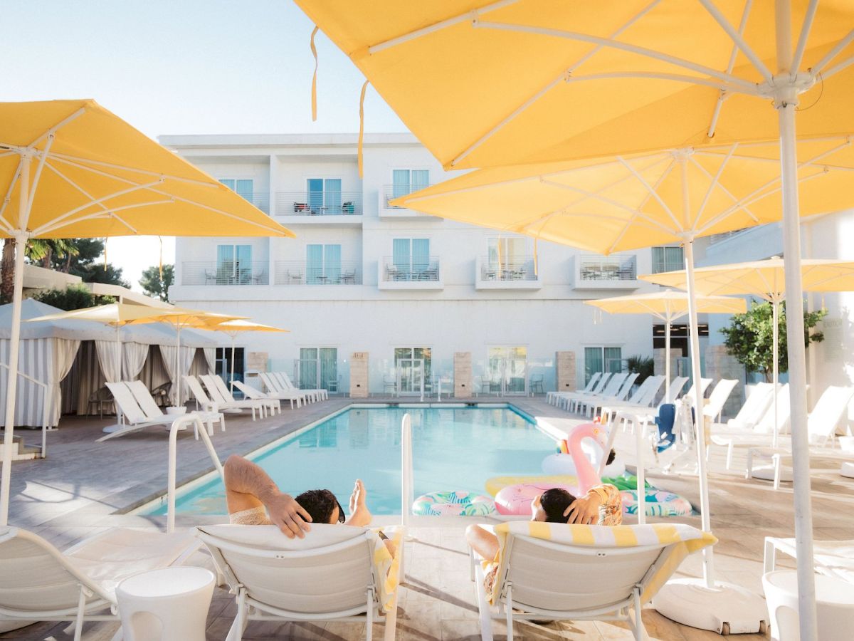A sunny poolside scene with white lounge chairs, yellow umbrellas, and a hotel building in the background.