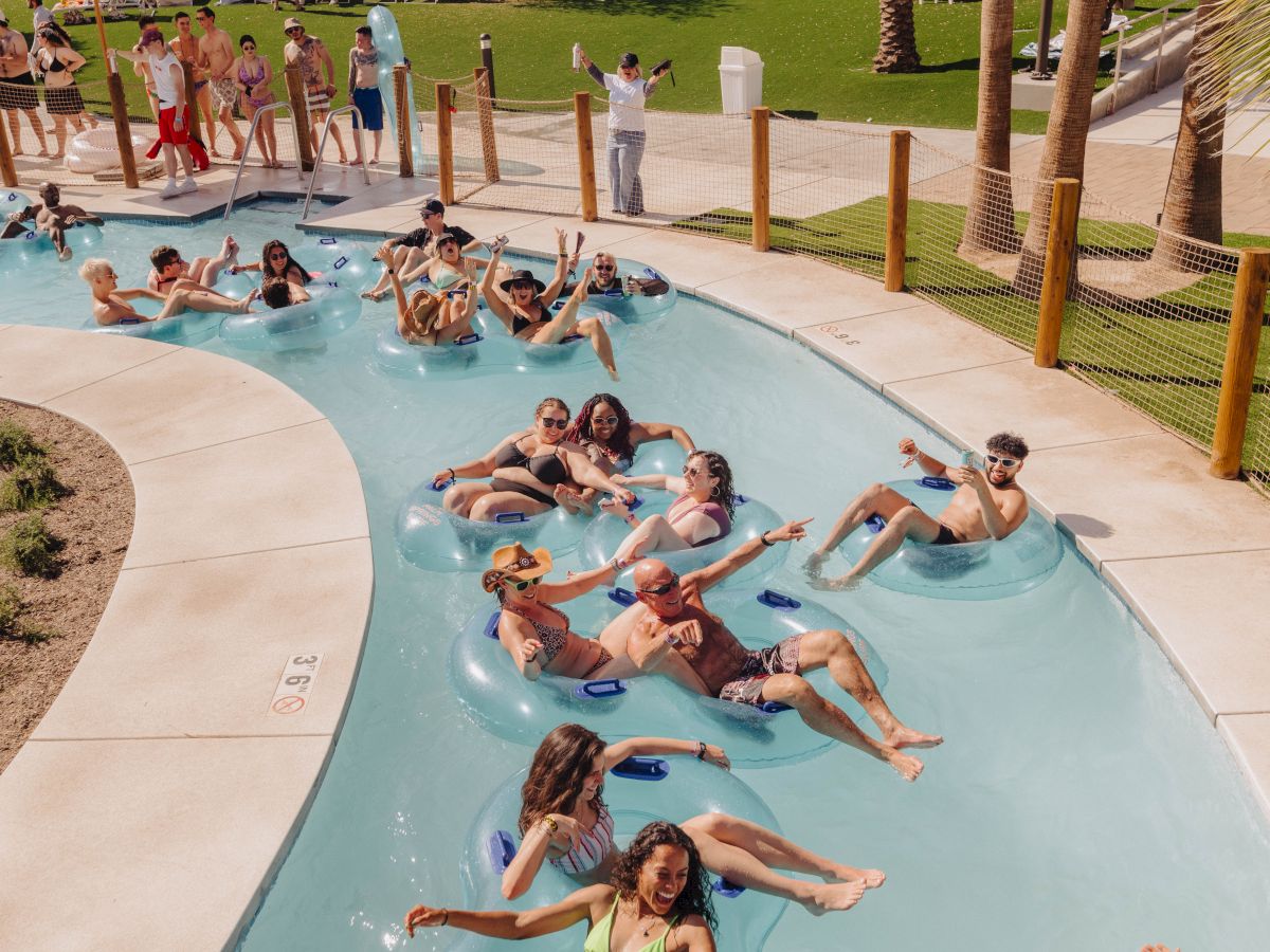 People relaxing on inflatable tubes in a lazy river under palm trees, with others in the background enjoying a sunny day by the pool.