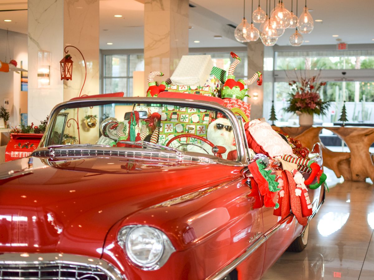 A shiny red vintage car decorated with Christmas garlands and ornaments indoors, with festive lights, trees, and holiday decor in the background.