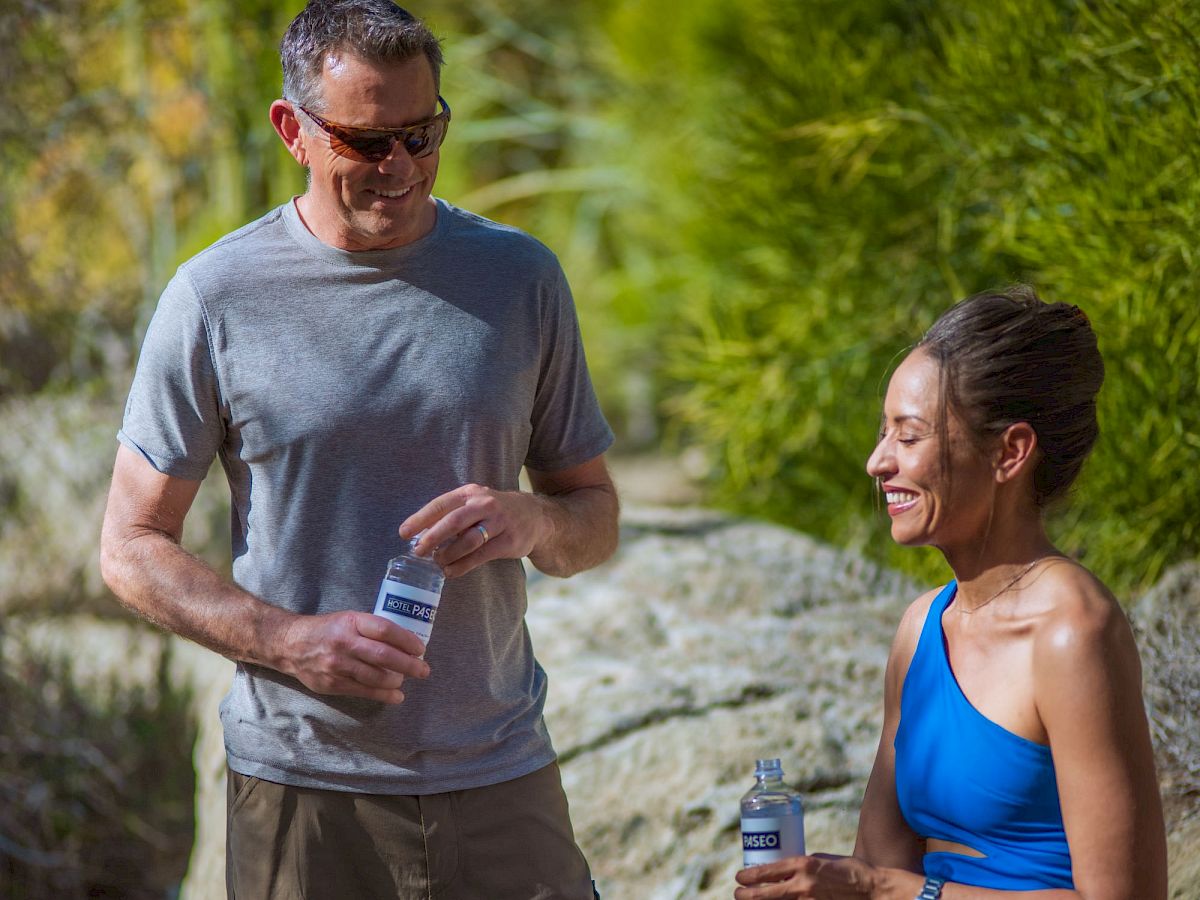 A man and woman in athletic gear chat and stretch after a run, each holding a water bottle, outdoors on a sunny trail.