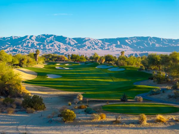A scenic golf course with green fairways, surrounded by desert landscape and distant mountains under a clear blue sky.