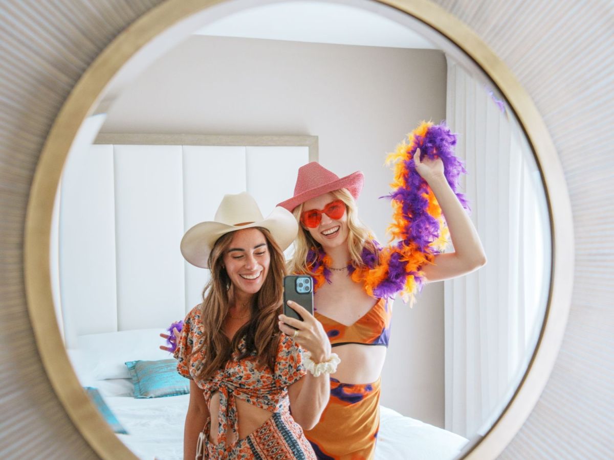 Two friends pose in a mirror selfie, wearing sun hats and colorful outfits while one holds a purple-orange boa, smiling brightly.