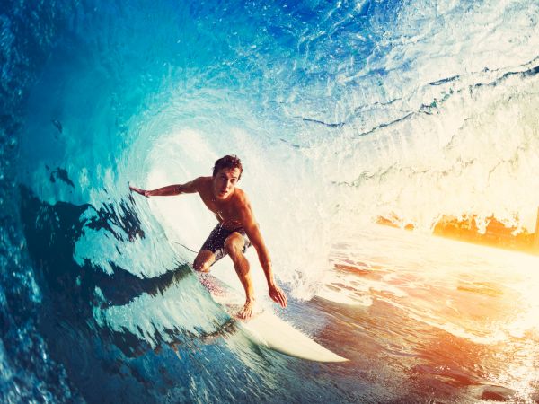 A surfer rides a curling ocean wave inside a bright, sunlit barrel, balancing on the board as blue water swirls around.