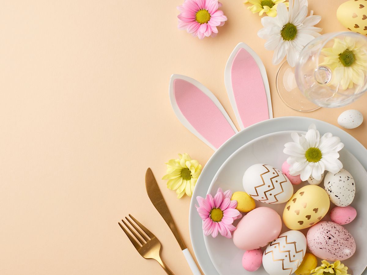 A festive Easter table: pastel eggs in a bowl, pink bunny ears, scattered flowers, and a gold knife and fork on a peachy background.