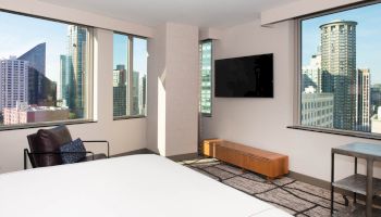 A modern hotel room with a bed, wall-mounted TV, chair, and large windows offering a cityscape view of tall buildings.