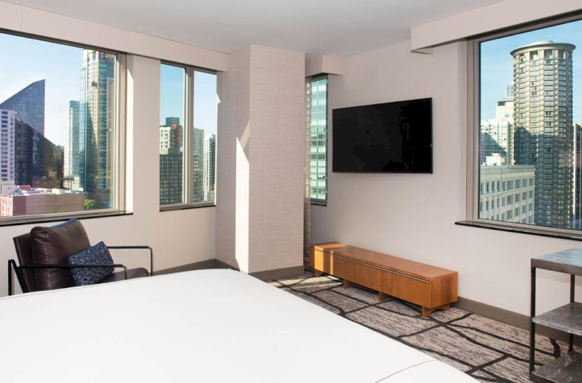 A modern hotel room with a bed, wall-mounted TV, chair, and large windows offering a cityscape view of tall buildings.