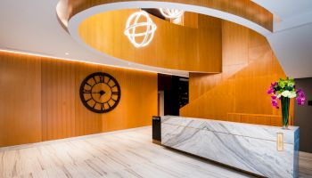 A modern lobby features a marble counter, wooden accents, a large wall clock, and decorative lighting fixtures, with flowers on the desk.
