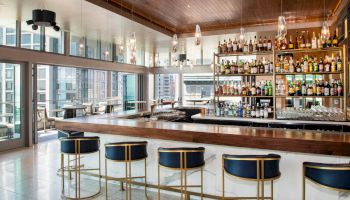 A modern bar with blue stools, a wooden counter, and shelves stocked with bottles, set in a bright room with city views.
