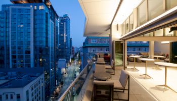 The image shows a modern cityscape with high-rise buildings and an outdoor terrace with seating, overlooking a street at dusk.