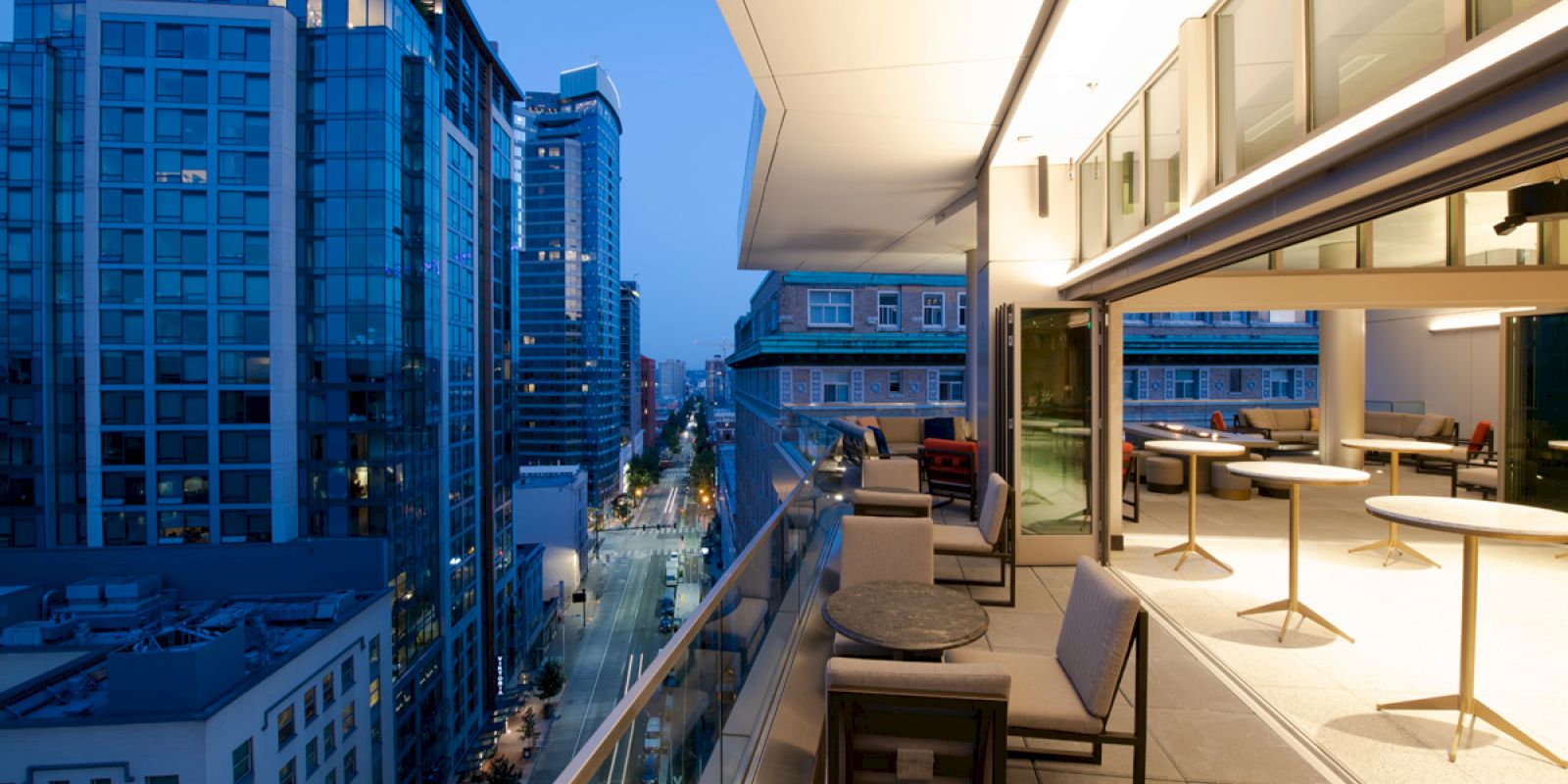 A modern rooftop terrace with seating overlooks an urban street lined with tall, illuminated buildings in the evening.