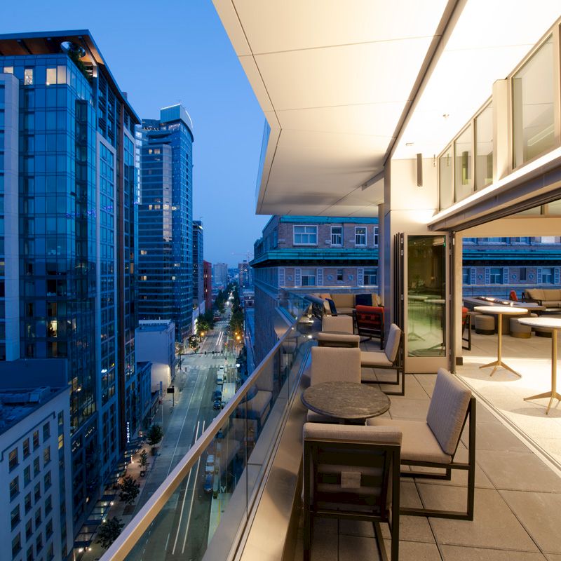 A modern balcony with tables and chairs overlooks a cityscape with tall buildings at dusk, under a clear blue sky.