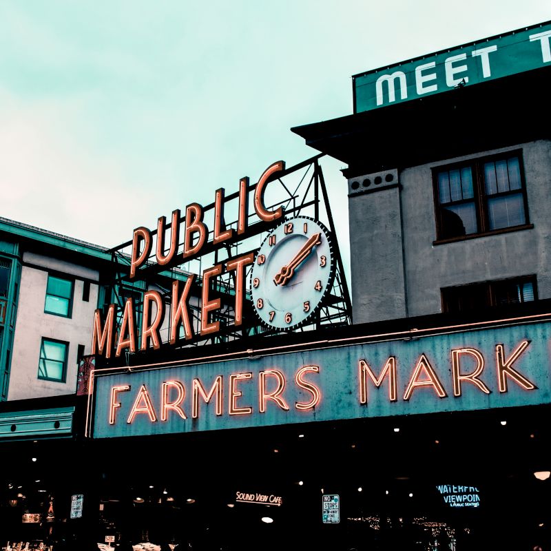 The image shows a neon sign for a public and farmers market, featuring a clock, against the backdrop of a building with more signage.