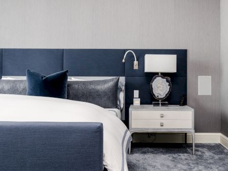 A modern bedroom with a navy headboard, white bedding, gray pillows, nightstand, lamp, and wall-mounted reading light.