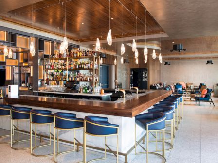 A modern bar with blue stools, hanging lights, and a stocked back bar, surrounded by a chic lounge area with seating.