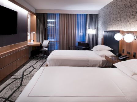 A hotel room with two beds, a wall-mounted TV, desk, chair, lamps, and patterned wallpaper.