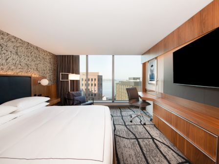 A modern hotel room with a large bed, desk, and a flat-screen TV. A window offers a city and water view, with stylish decor elements.