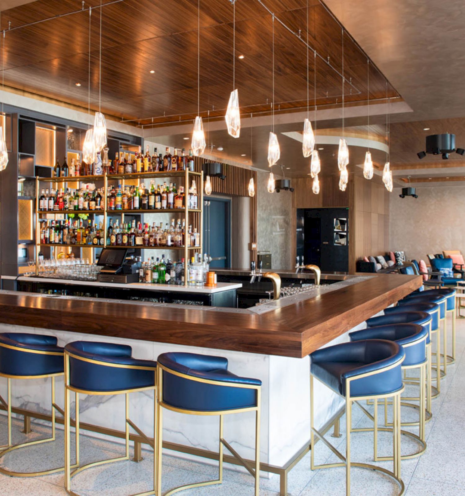 A modern bar with blue chairs, wooden counters, and hanging lights. It features a wide selection of bottles and a stylish seating area.
