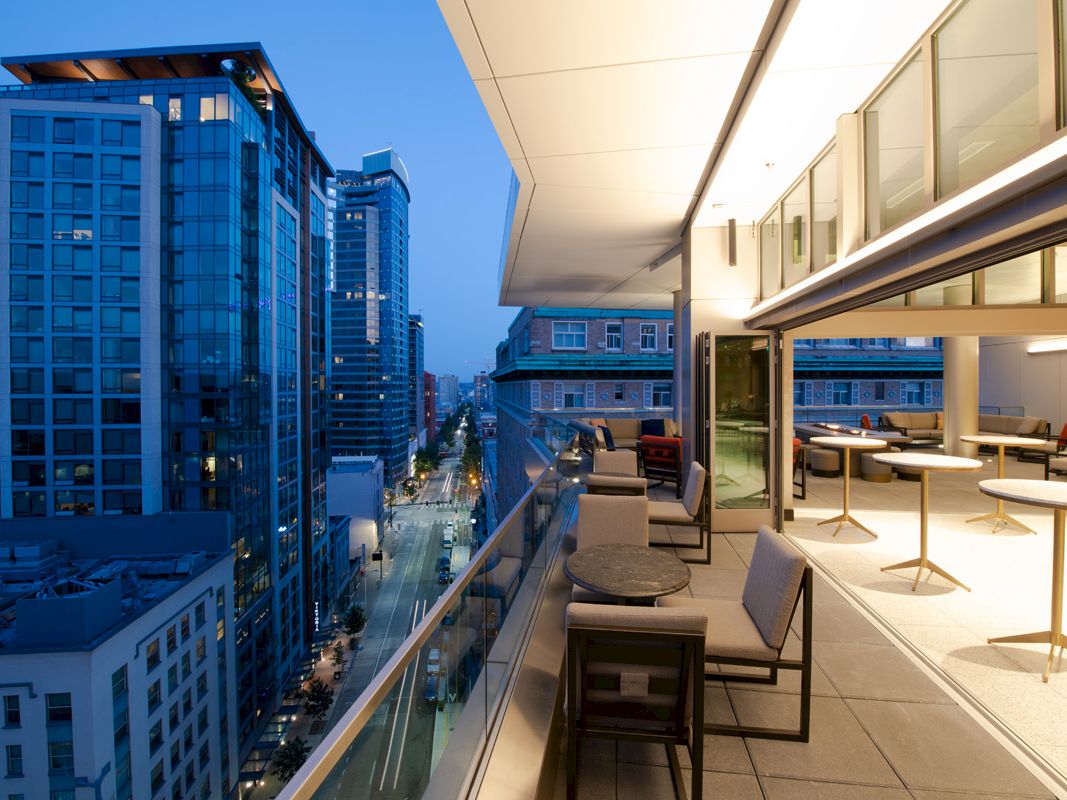 A modern rooftop terrace overlooking a cityscape with tall buildings, featuring seating and tables under warm lighting.