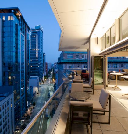 A modern rooftop patio with seating overlooks a cityscape with tall buildings and a street, all under a clear evening sky.