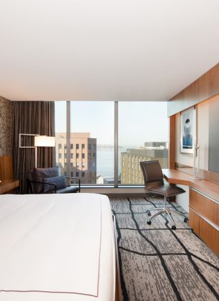 A modern hotel room with a large bed, desk, chair, and wall-mounted TV, featuring a city view through large windows.