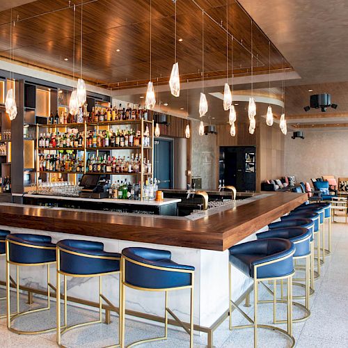 This image shows a stylish bar with hanging lights, blue bar stools, a well-stocked drink shelf, and a seating area with couches.