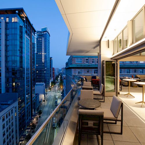 The image shows a modern urban balcony with seating, overlooking a cityscape with tall buildings and illuminated streets at dusk.
