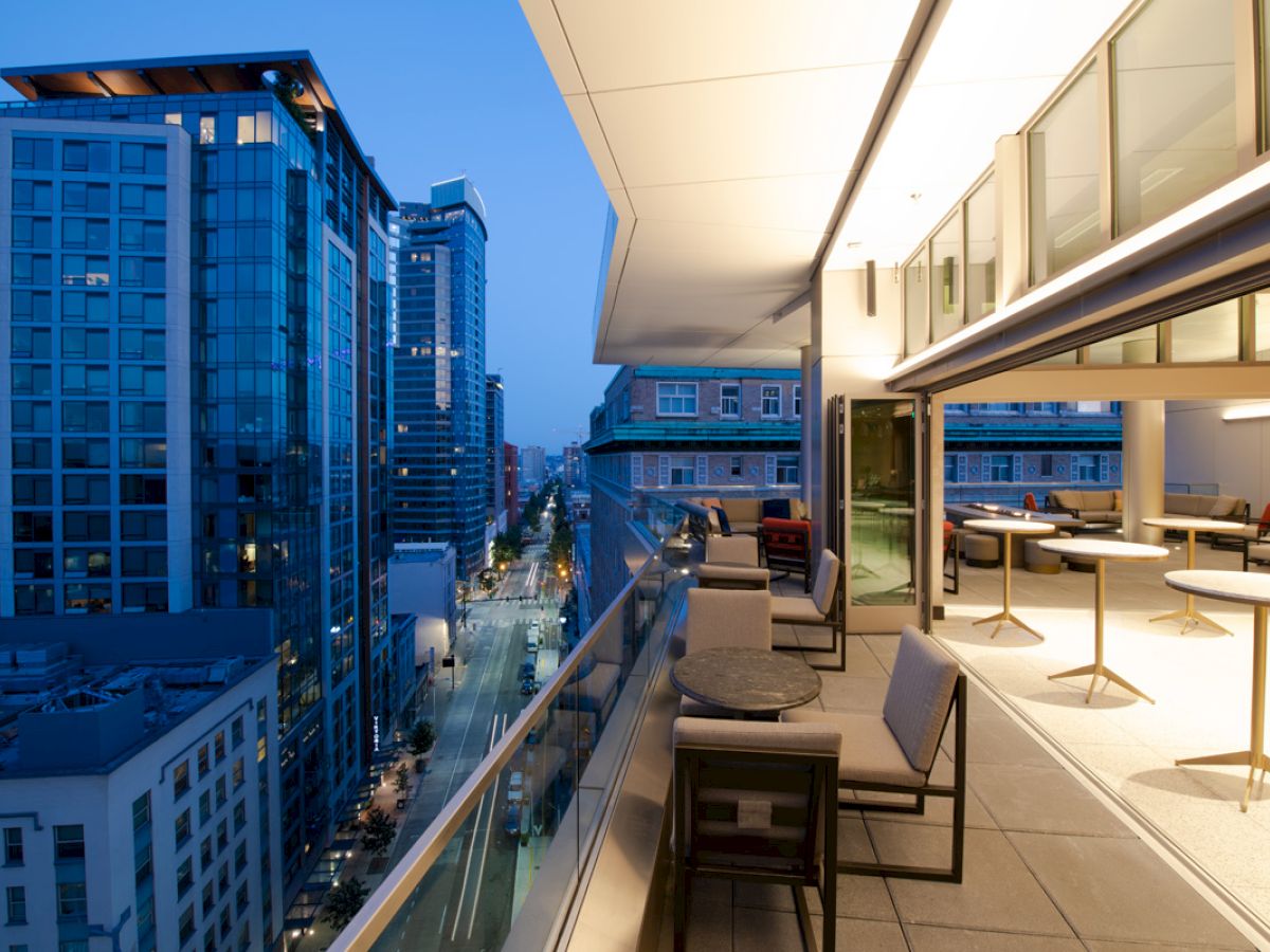 A modern cityscape at twilight, with tall buildings and a sleek balcony featuring outdoor seating and tables, extending into the scene.