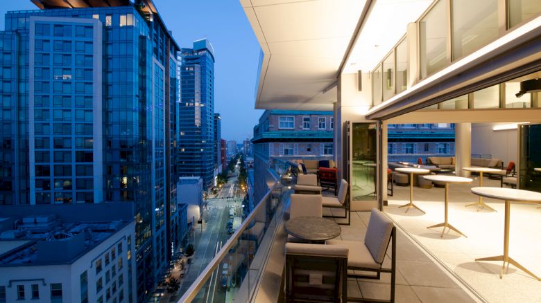 A modern cityscape at twilight, with tall buildings and a sleek balcony featuring outdoor seating and tables, extending into the scene.