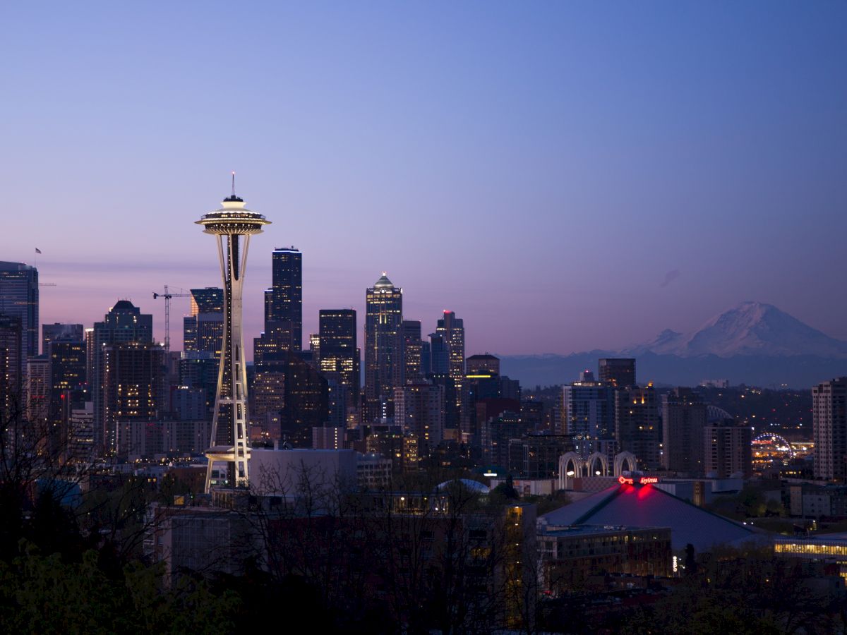 Seattle skyline at dusk featuring the Space Needle, city buildings, and Mount Rainier in the background, under a purple-blue sky.