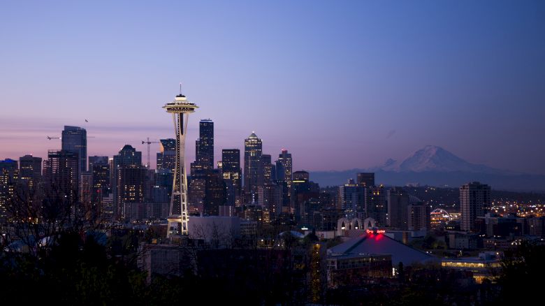 Seattle skyline at dusk featuring the Space Needle, city buildings, and Mount Rainier in the background, under a purple-blue sky.